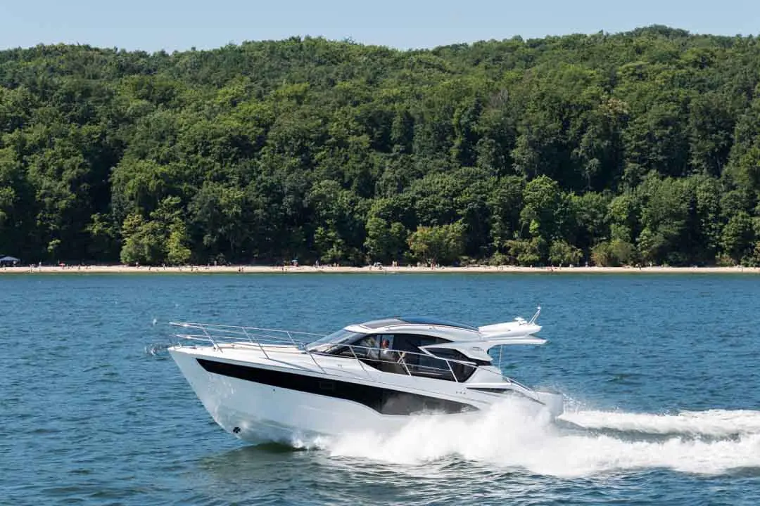 A sleek white motorboat glides across clear blue water near a green forest and sandy beach, with people sunbathing in the distance.