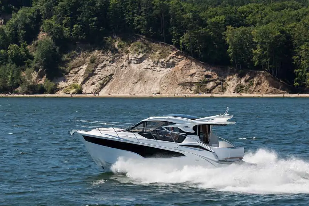 A sleek white motorboat speeds across a clear blue lake, surrounded by lush green hills and a sandy beach in the background.