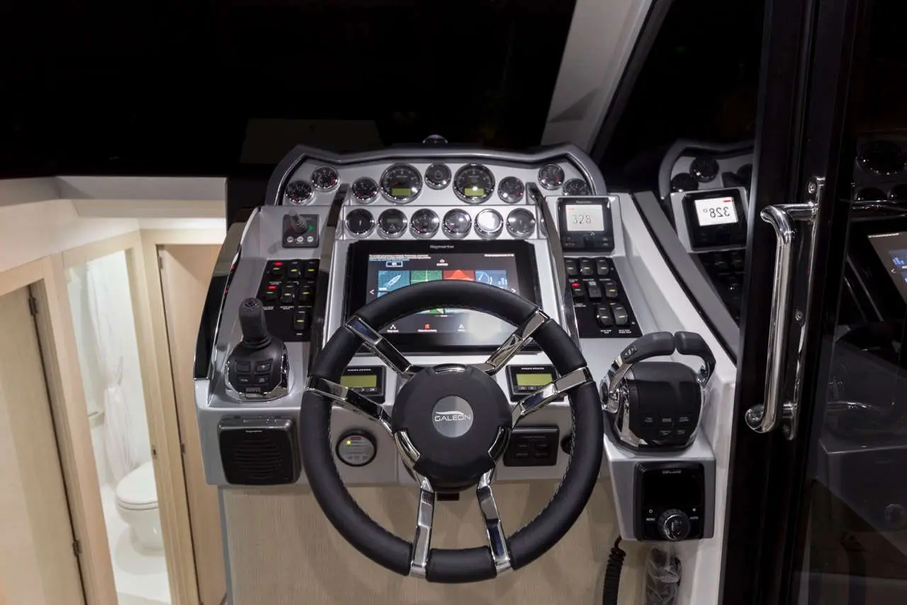 Luxury boat steering wheel and control panel with digital displays, gauges, and buttons; cabin visible in the background.