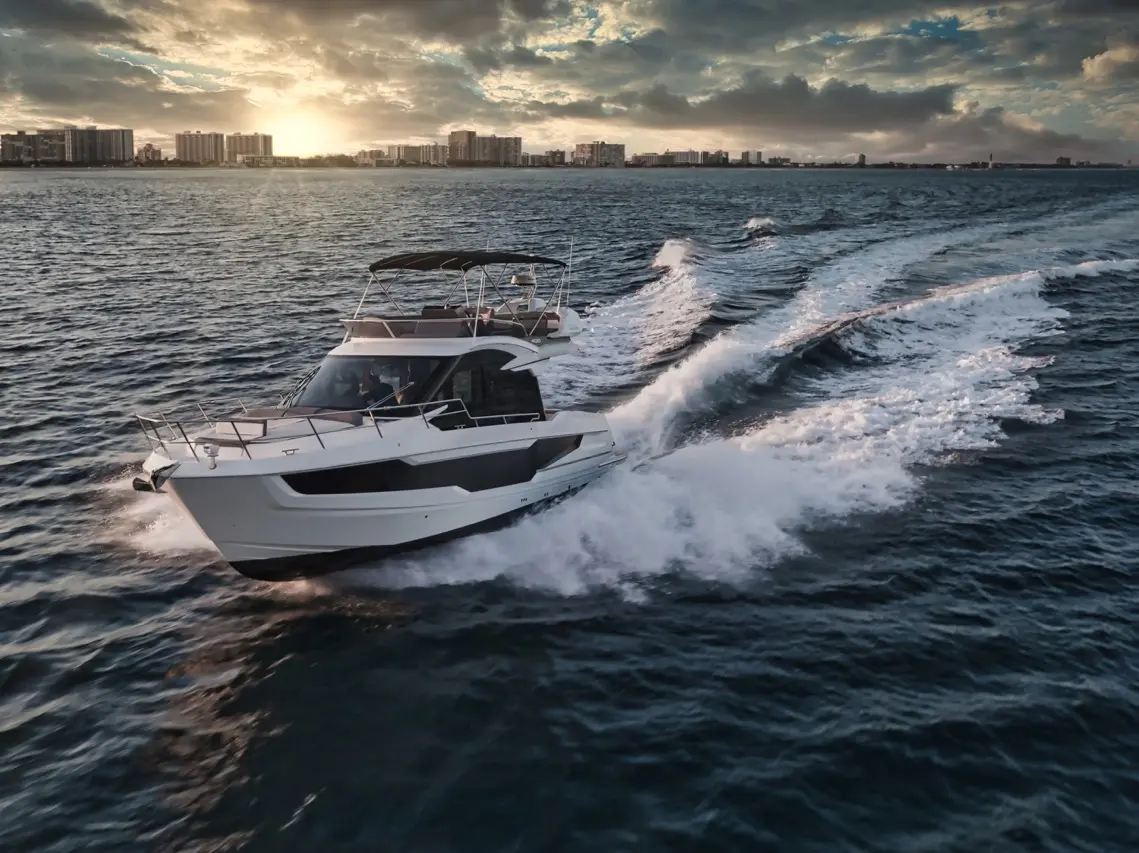 A modern white yacht speeds through the water, creating waves as the sun sets behind a skyline.