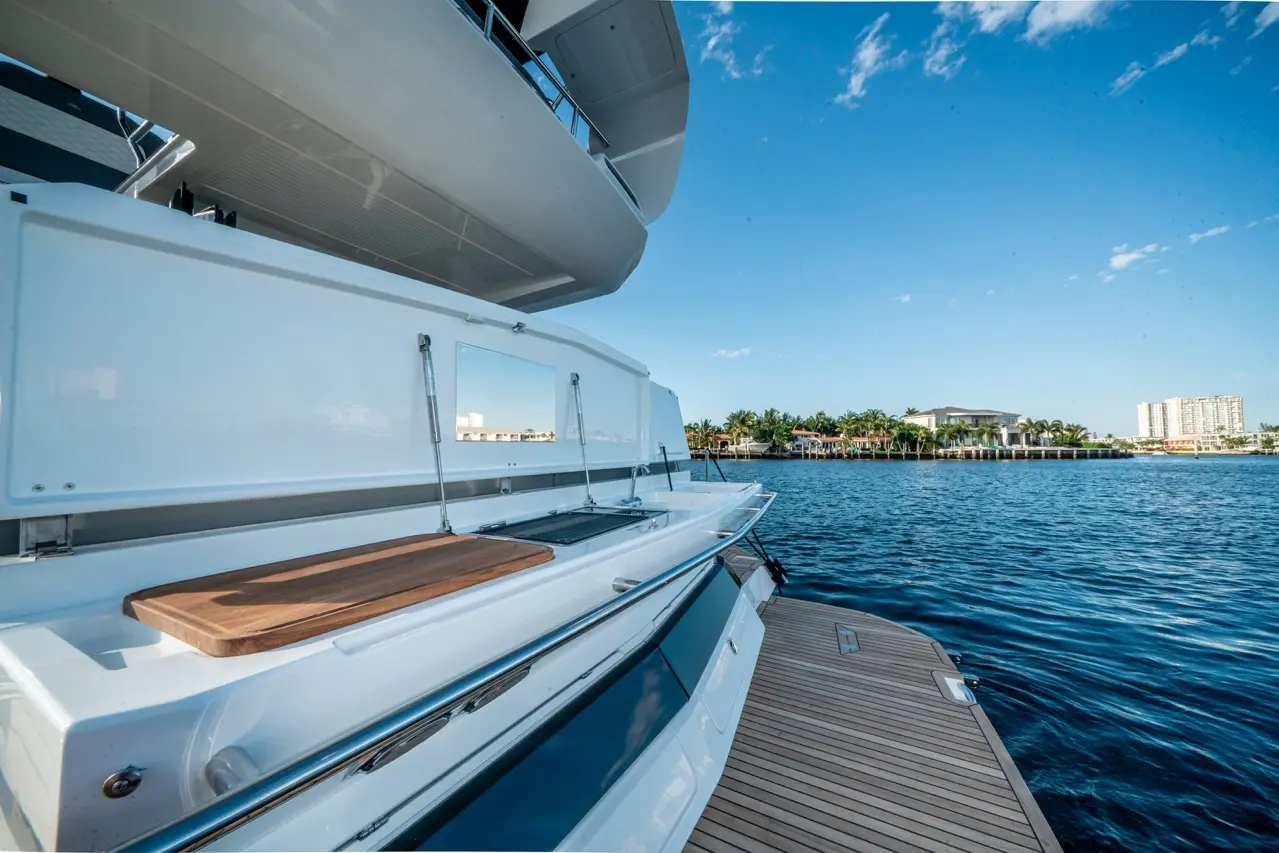 Luxury yacht with a wooden serving surface docked by calm waters, featuring scenic waterfront homes and a clear blue sky.