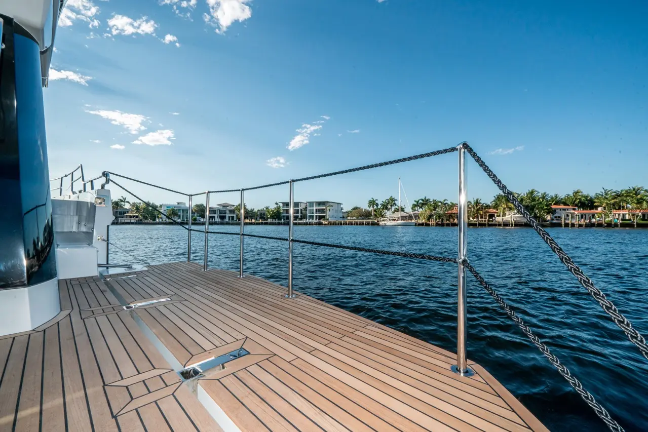 View from a yacht deck showcasing wooden flooring, metal railings, and a scenic waterfront with luxury homes and palm trees.