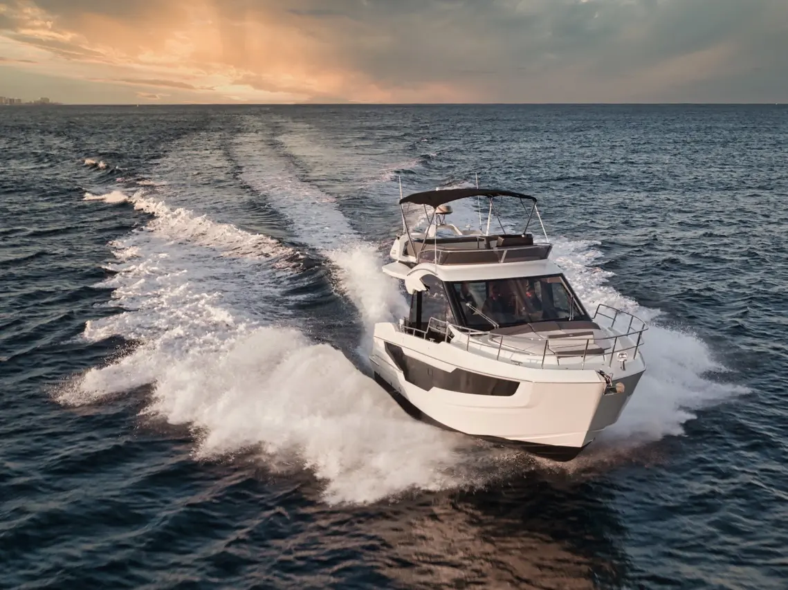 A modern motorboat speeds across the ocean, leaving a wake behind it, under a sunset sky with scattered clouds.