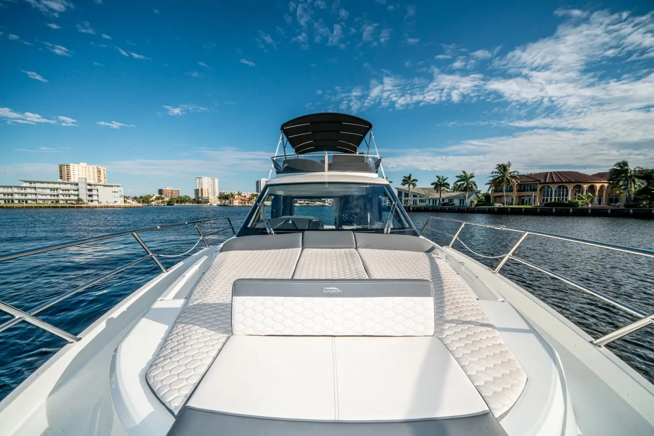 View from the bow of a luxury boat with a patterned seating area, water, palm trees, and modern buildings in the background.