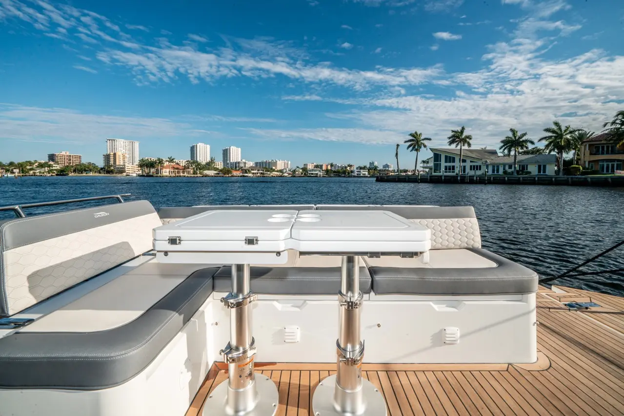 View from a boat showing a white table, cushioned seating, and waterfront homes with palm trees under a blue sky.