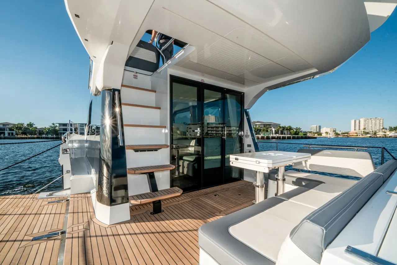 Modern yacht interior with stairs, glass doors, seating area, and views of waterfront properties under a clear blue sky.