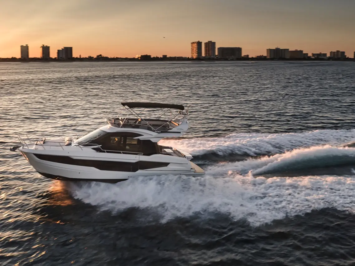 A sleek white motor yacht speeds across calm waters at sunset, with city buildings visible in the background.