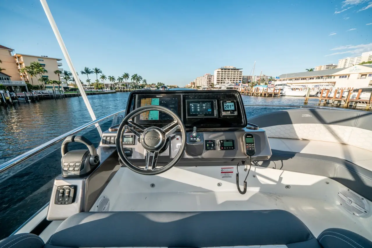 Boat cockpit view with steering wheel, digital screens showing navigation data, and waterfront scenery in the background.
