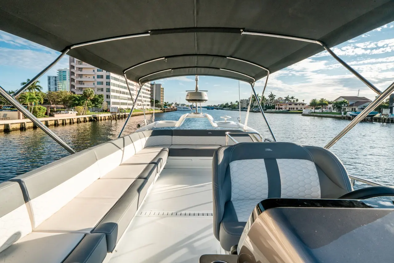Interior view of a boat with padded seating, overlooking a waterway lined with buildings and palm trees under a blue sky.