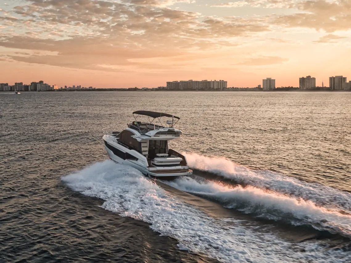 A luxury motorboat speeds through the water at sunset, leaving a white wake against the serene ocean and city skyline.