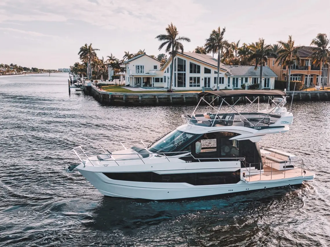 Luxury motor yacht cruising along a waterfront with palm trees and modern homes in the background.