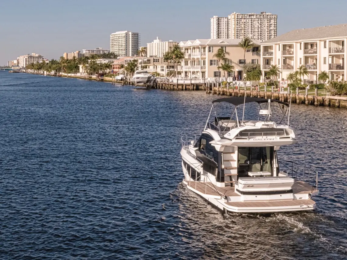 A sleek yacht navigates a calm waterway lined with modern buildings and palm trees under a clear sky.