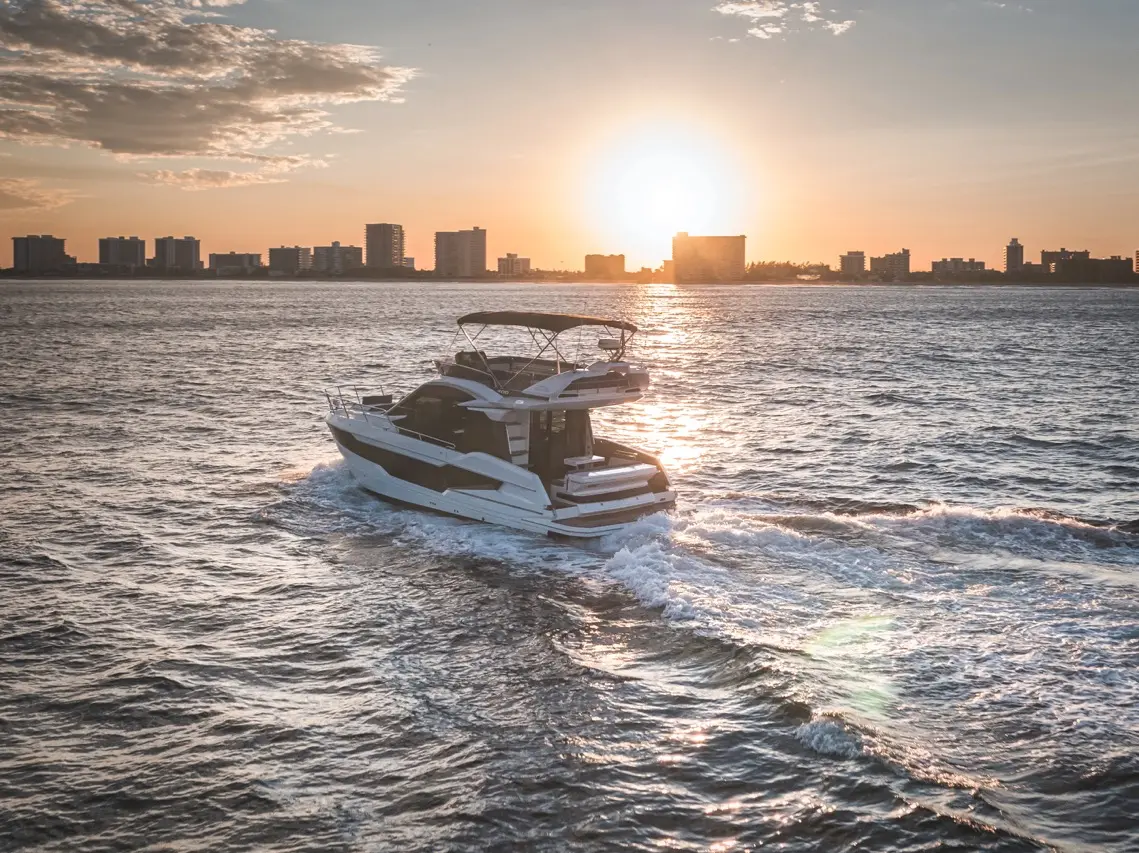 A luxury motor yacht glides over shimmering waters at sunset, with a city skyline silhouetted in the background.