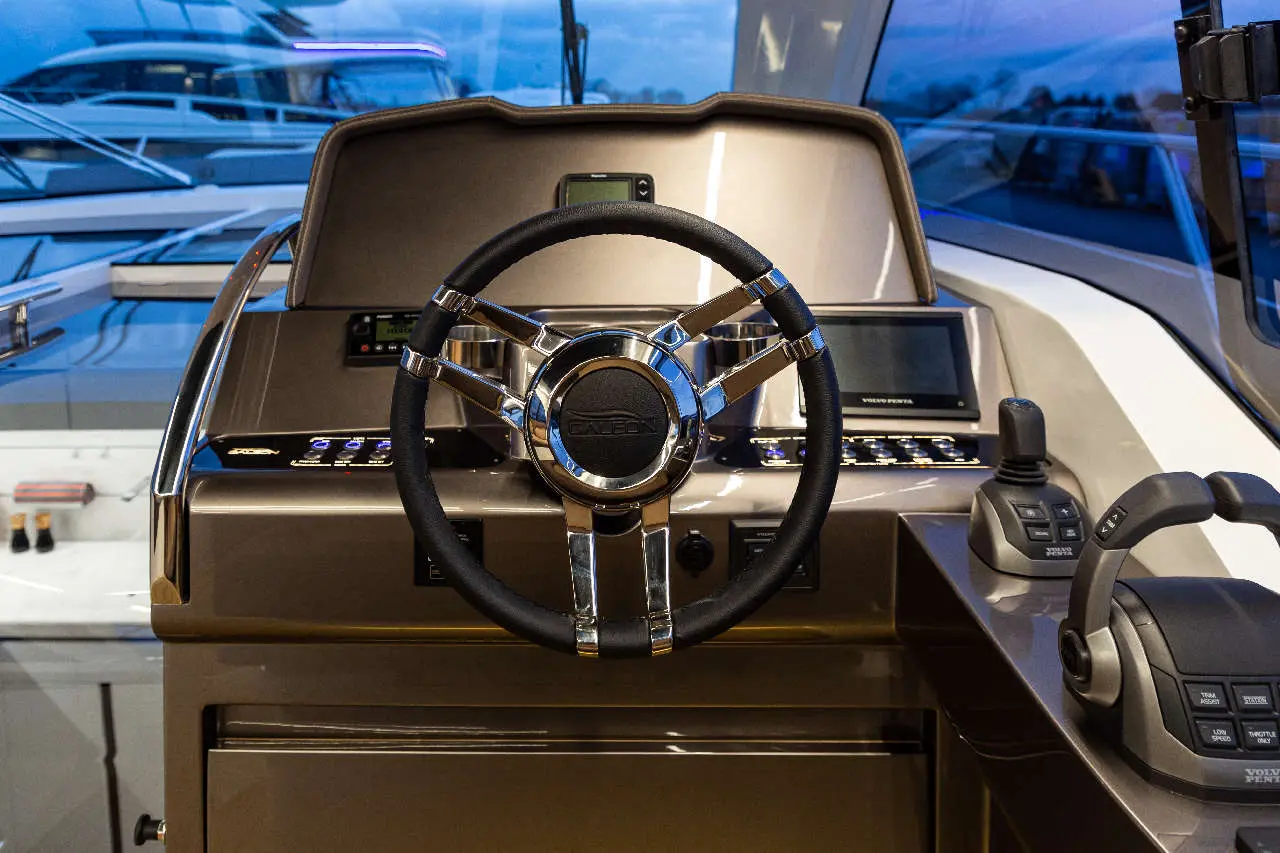 Modern boat control panel featuring a chrome steering wheel, digital display, and navigation controls, with boats in the background.