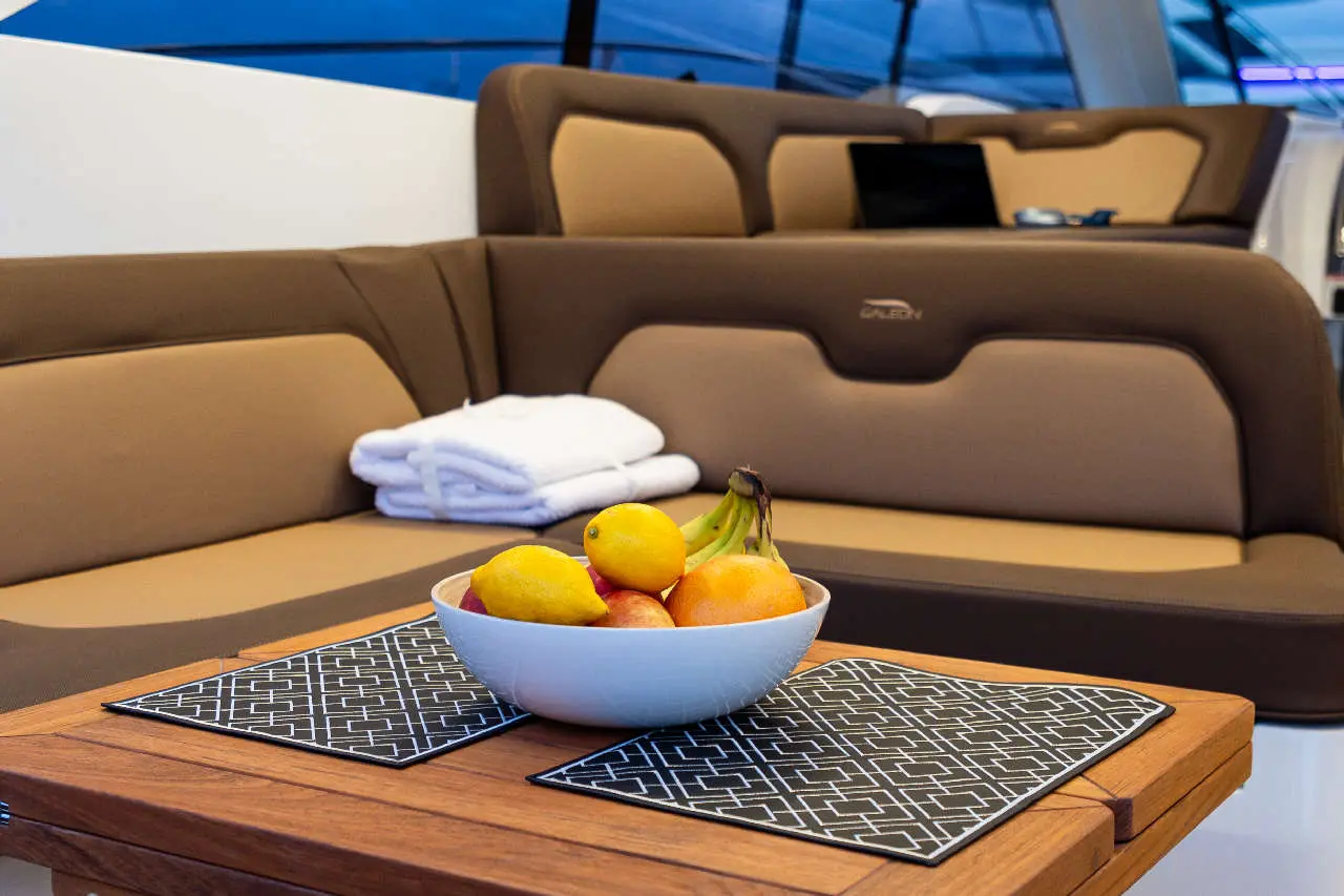 Bowl of mixed fruit on a wooden table with placemats, set in a luxurious yacht interior featuring brown seating.