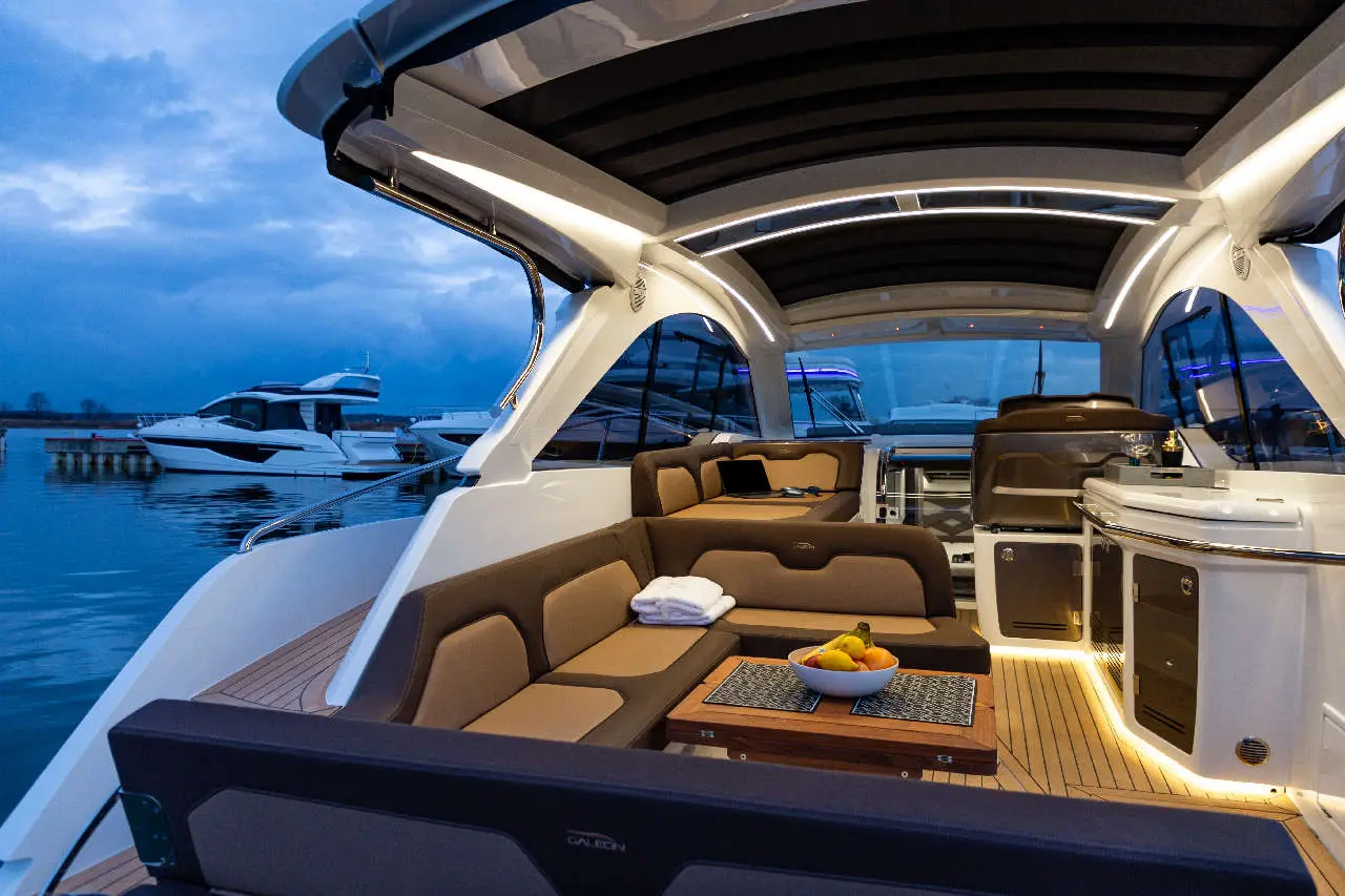 Luxury boat interior with seating, a fruit bowl, and evening lighting, overlooking parked boats on calm water.