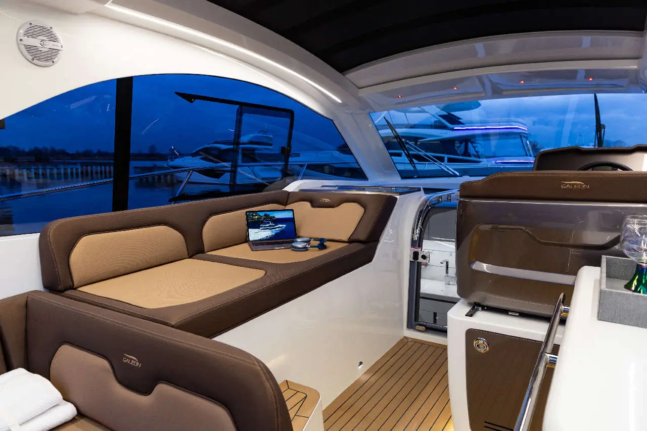 Modern boat interior with plush seating, a laptop on a table, and views of the water and other boats. Galeon branding visible.