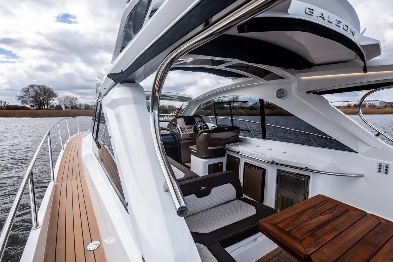 Luxury yacht side view showing a teak deck, modern cockpit, and soft seating, with water and trees in the background.