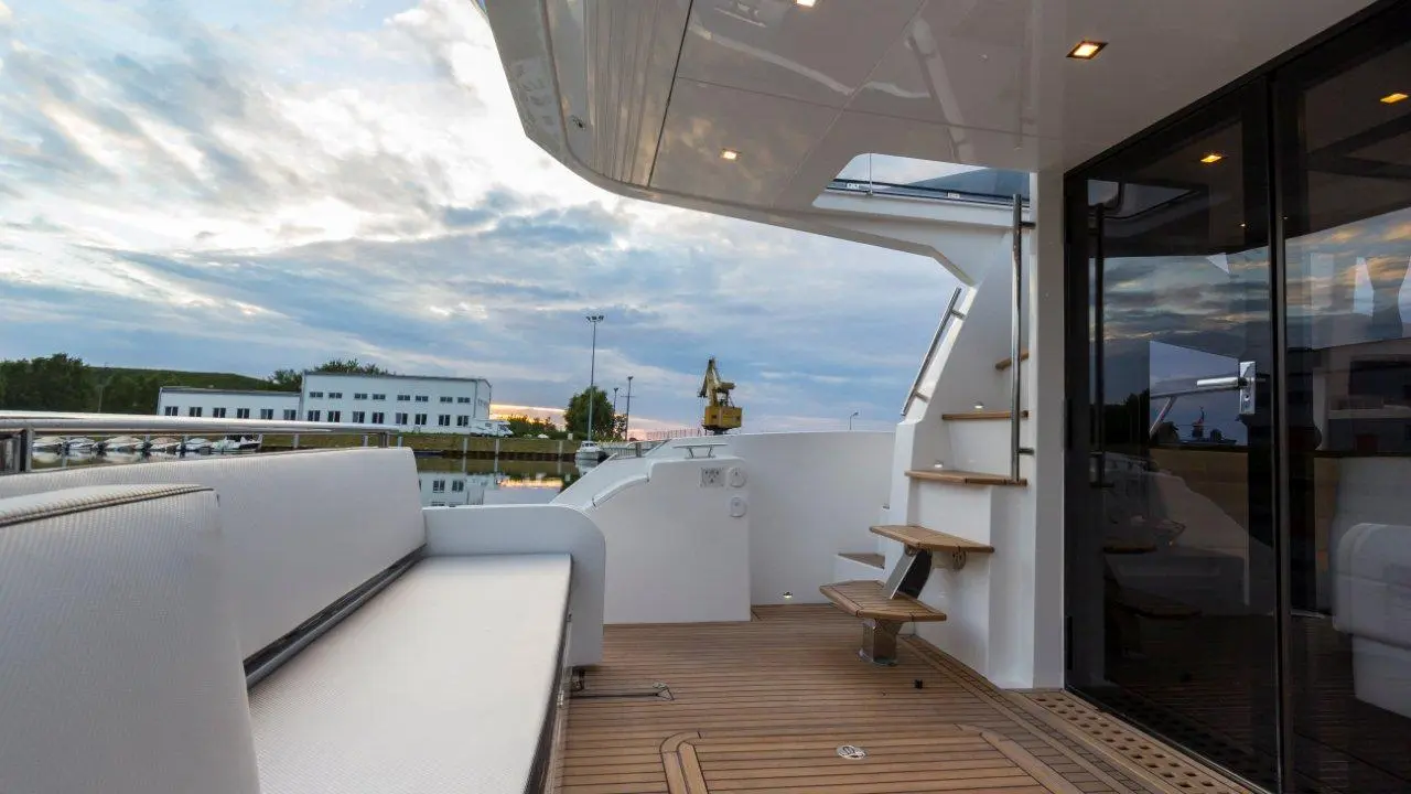Luxury yacht deck with white seating, wooden steps, and glass doors, overlooking a marina and cloudy sky at sunset.