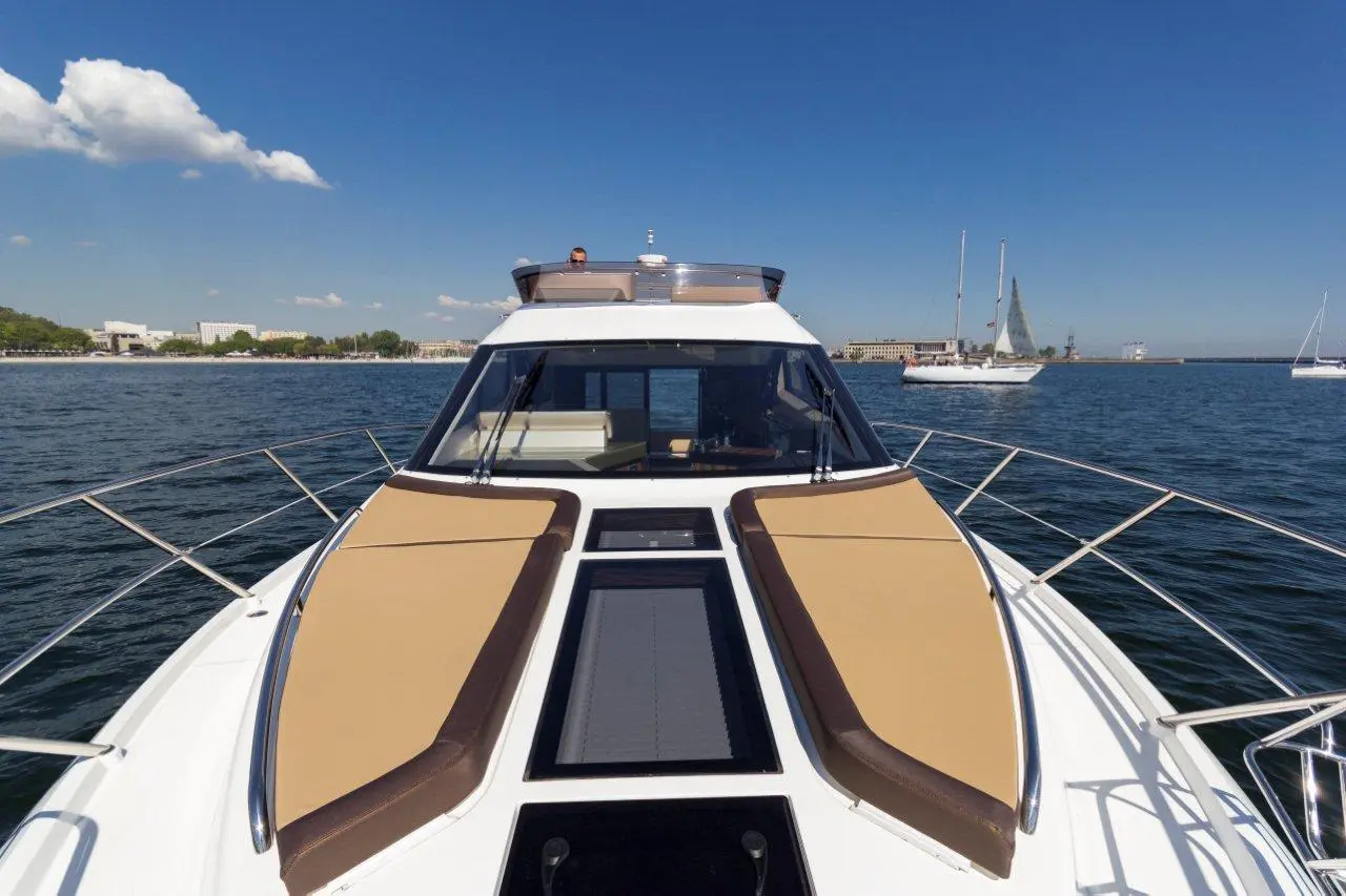 Deck view of a luxury yacht on calm water, featuring tan seating, a clear blue sky, and distant sailboats in the background.