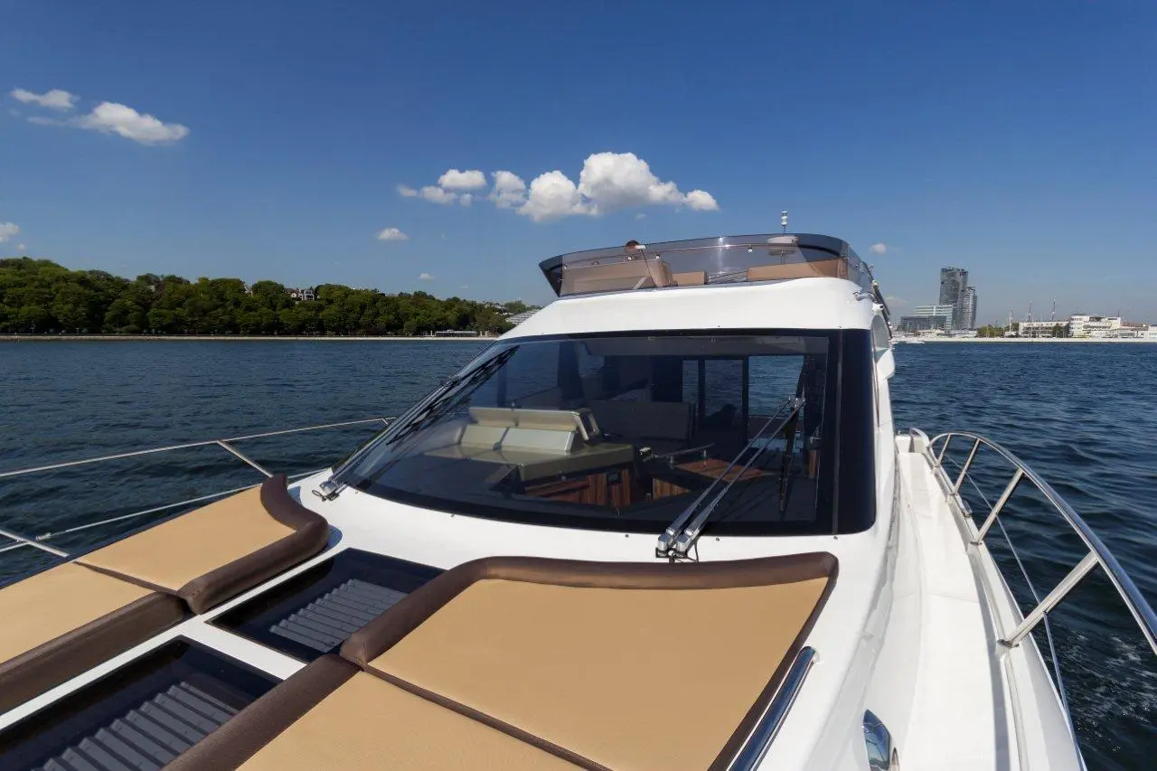Luxury yacht with sunbeds, waterfront view, and city skyline under a clear blue sky and scattered clouds.