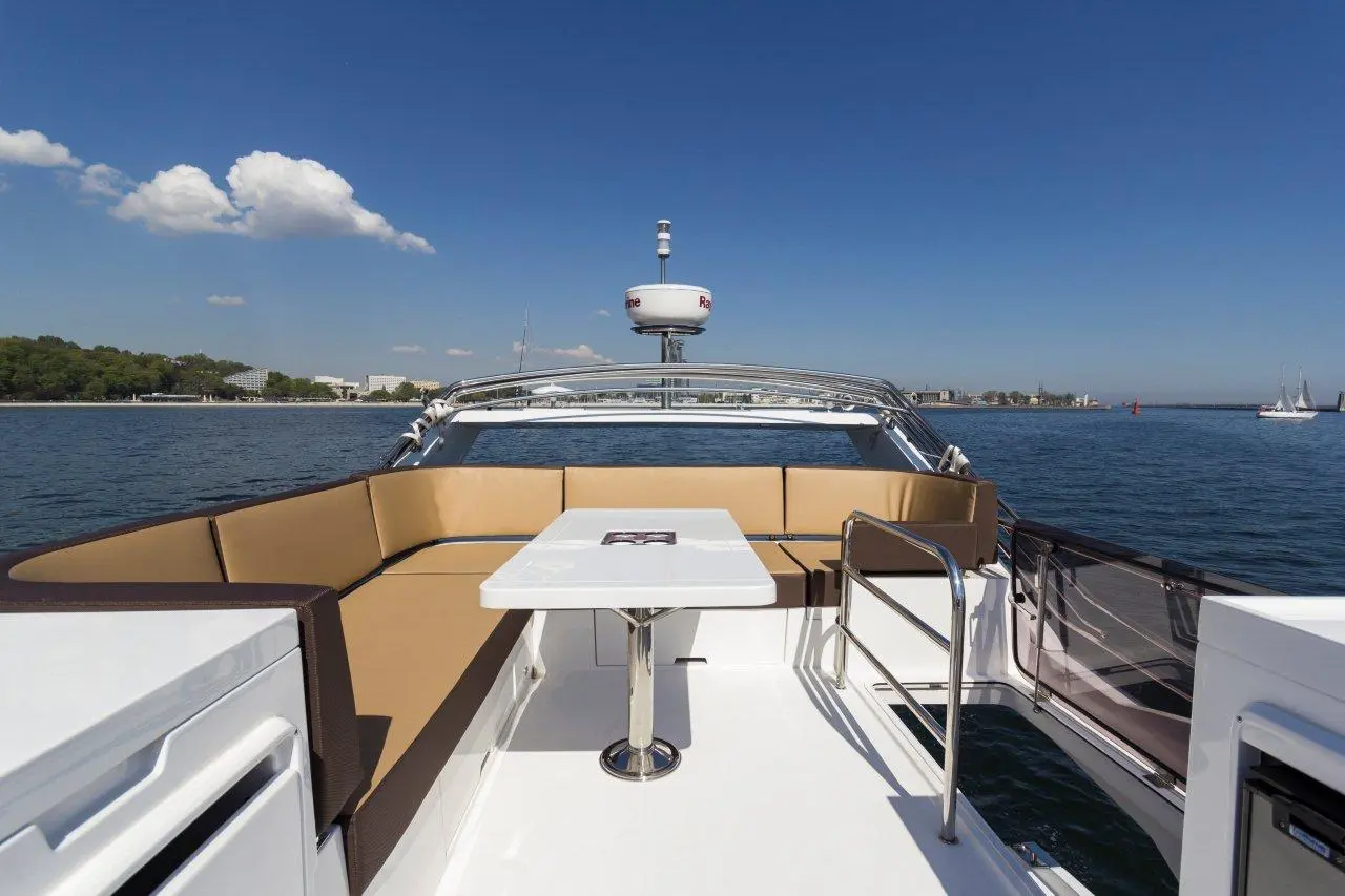 View from a yacht's bow featuring beige cushions, a white table, and a blue sky with clouds over calm waters and shoreline.