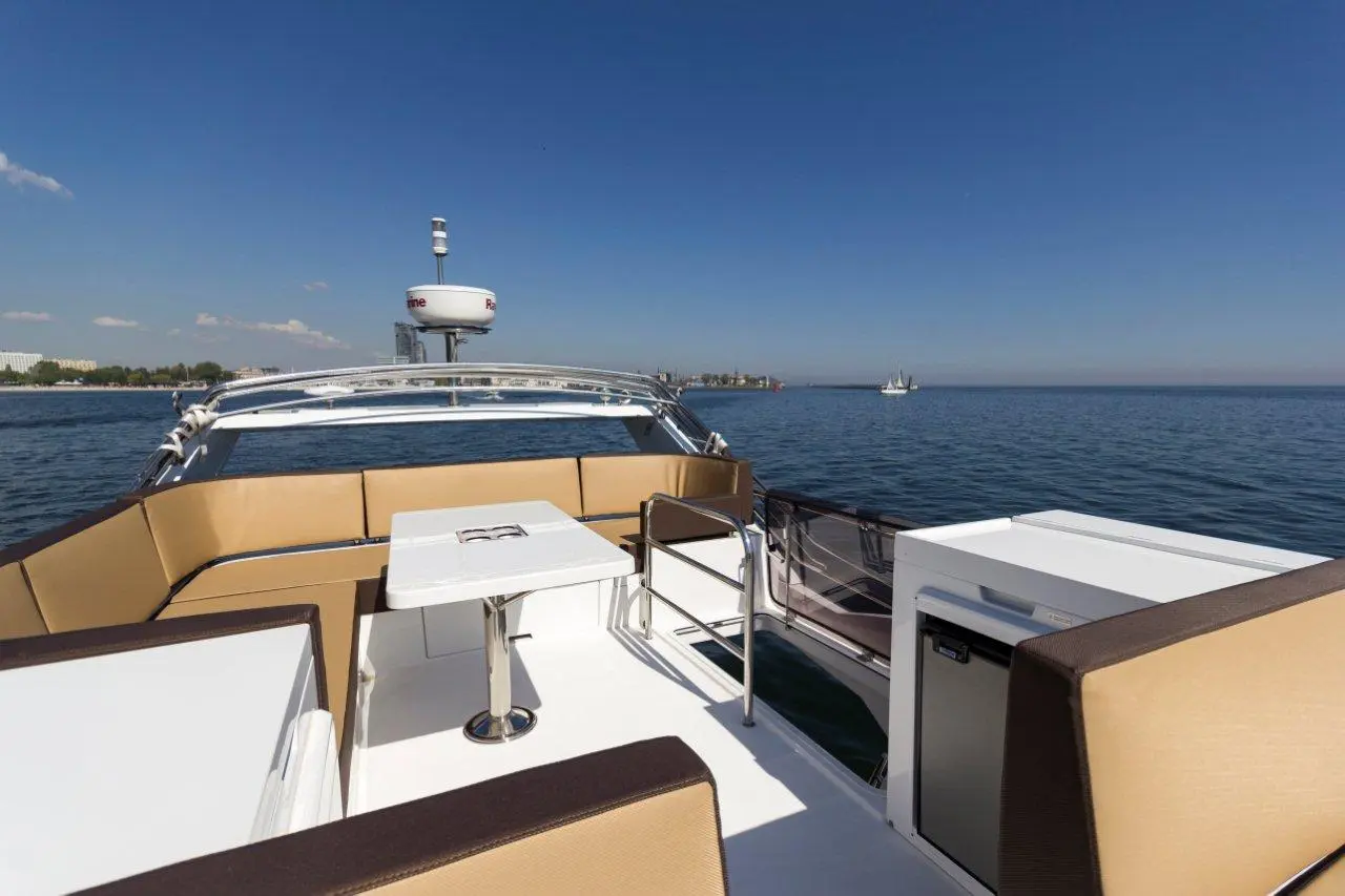 View from a boat's deck, featuring cushioned seating and a table, with a serene water landscape and clear blue sky.