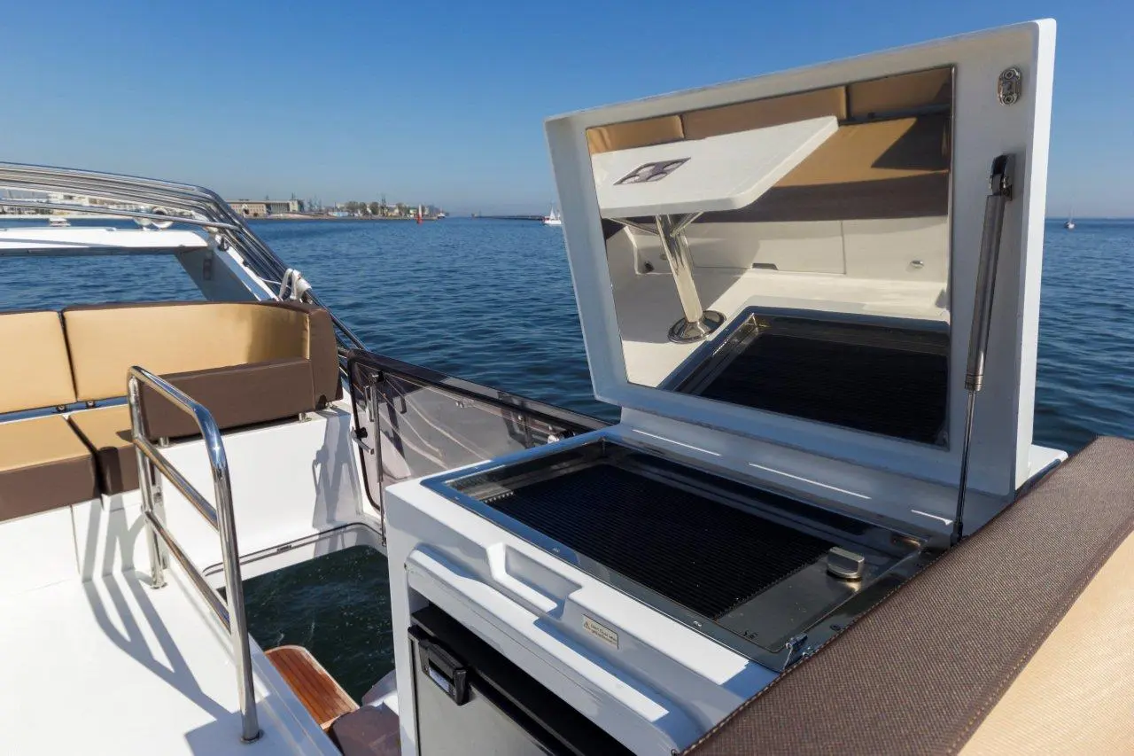A close-up of a boat's deck featuring a grill with lid open, overlooking calm water and distant shoreline under clear blue sky.