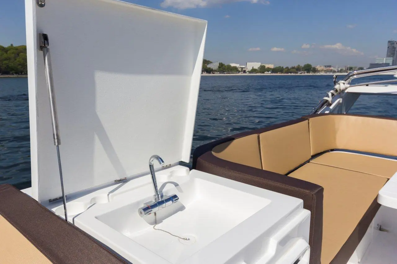 Boat cockpit with a sink and faucet under a lift-up lid, surrounded by cushioned seating, with water visible in the background.