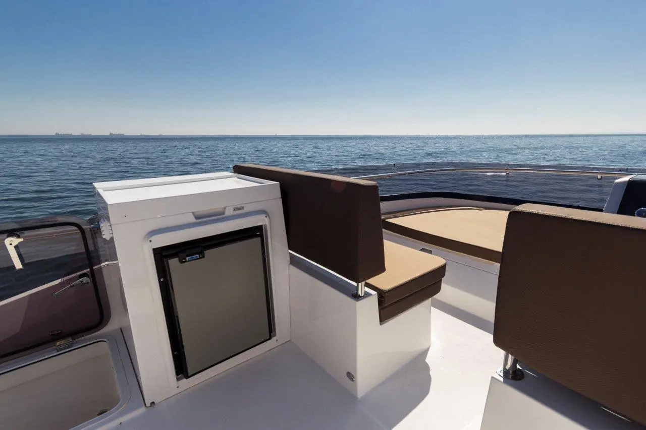 Modern boat seating with brown cushions, a small fridge, and a view of calm waters under a clear blue sky.