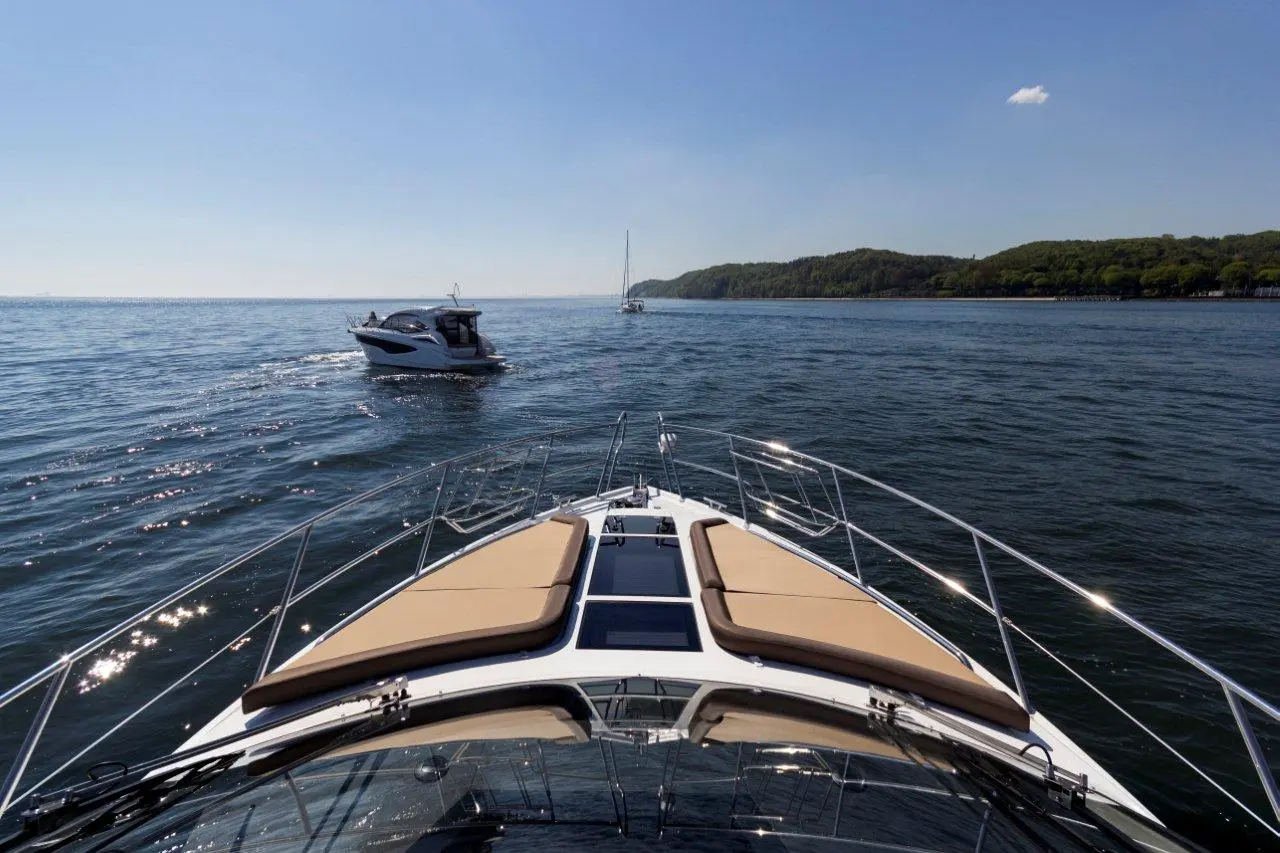 A motorboat navigates on calm water, with a scenic shoreline and another boat in the distance under a clear blue sky.
