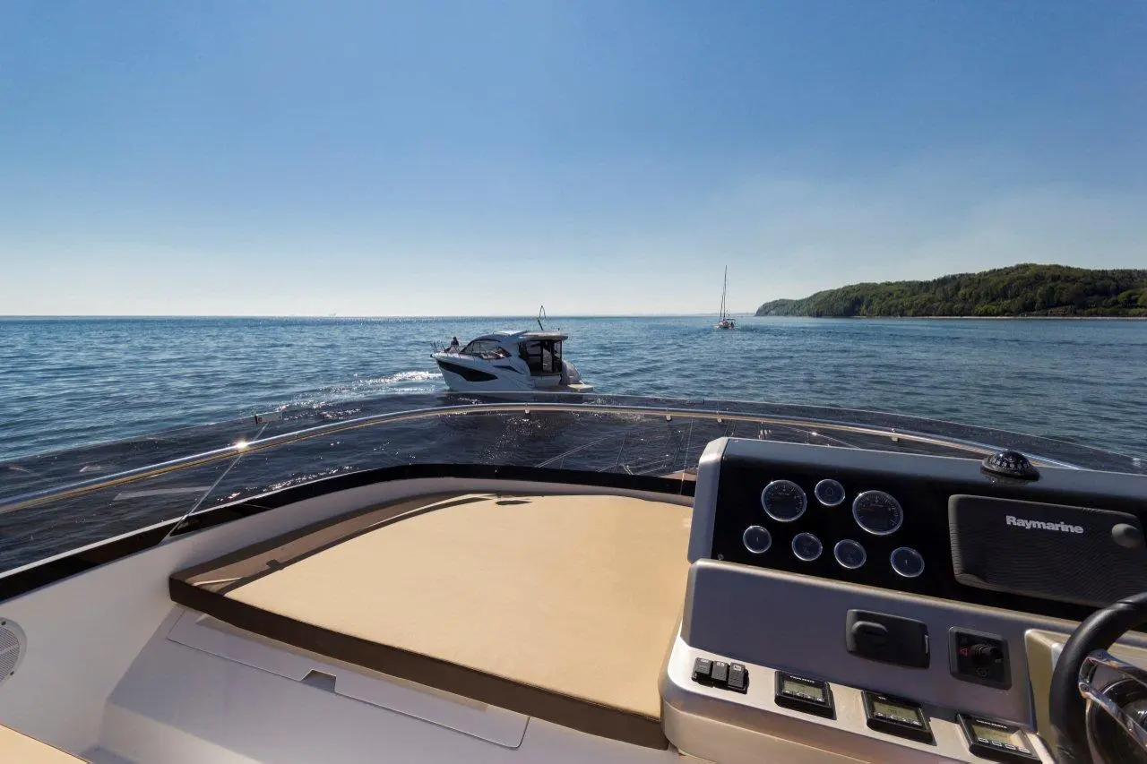 View from the helm of a boat, showing water, another boat, and distant shoreline under a clear blue sky.