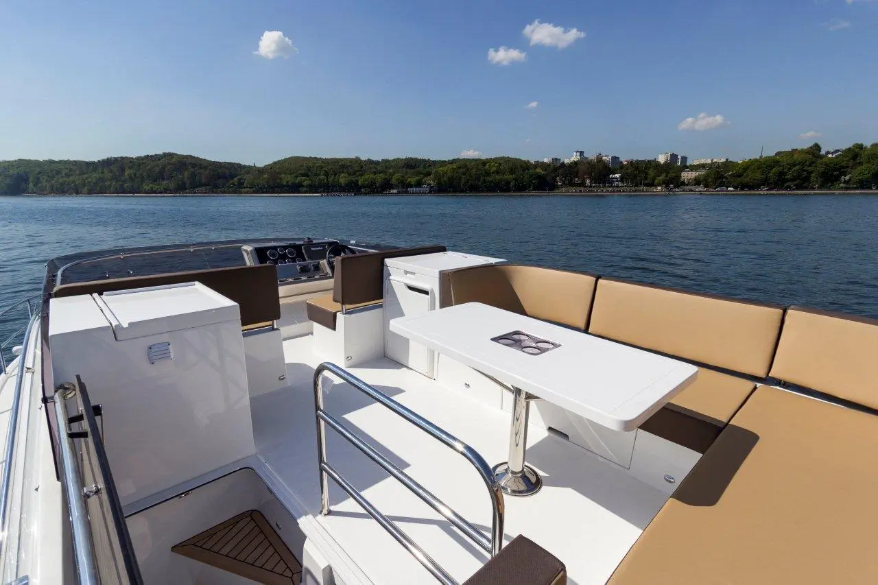 Brightly lit boat deck with tan cushions, a white table, and a view of calm water and green hills under a blue sky.