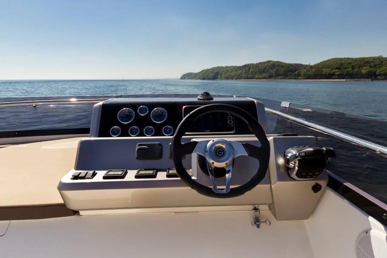 Boat's dashboard with steering wheel, gauges, buttons, and scenic view of water and landscape under clear skies.