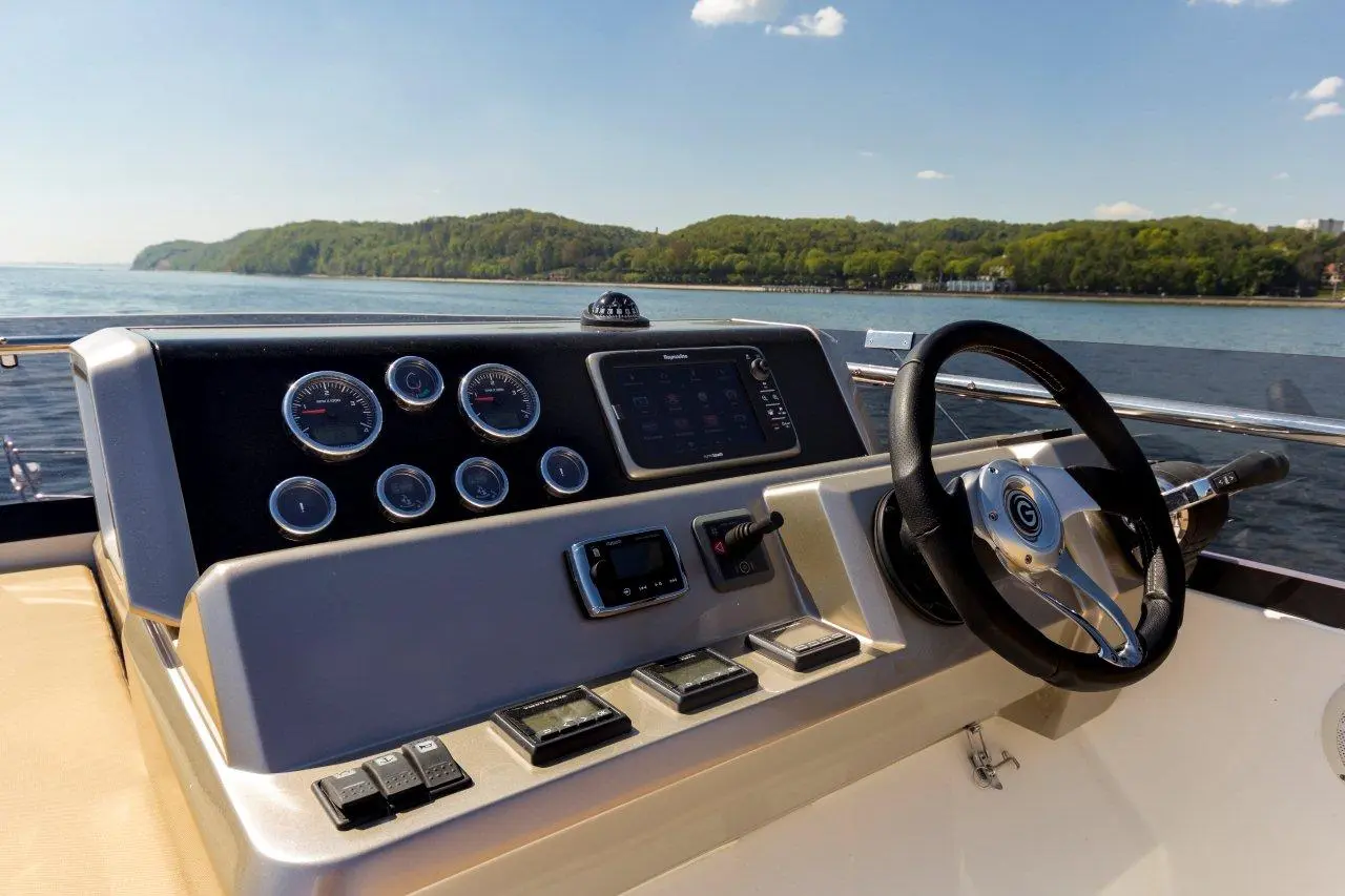 Boat cockpit dashboard with gauges, controls, and a steering wheel overlooking a scenic water view and green hills.