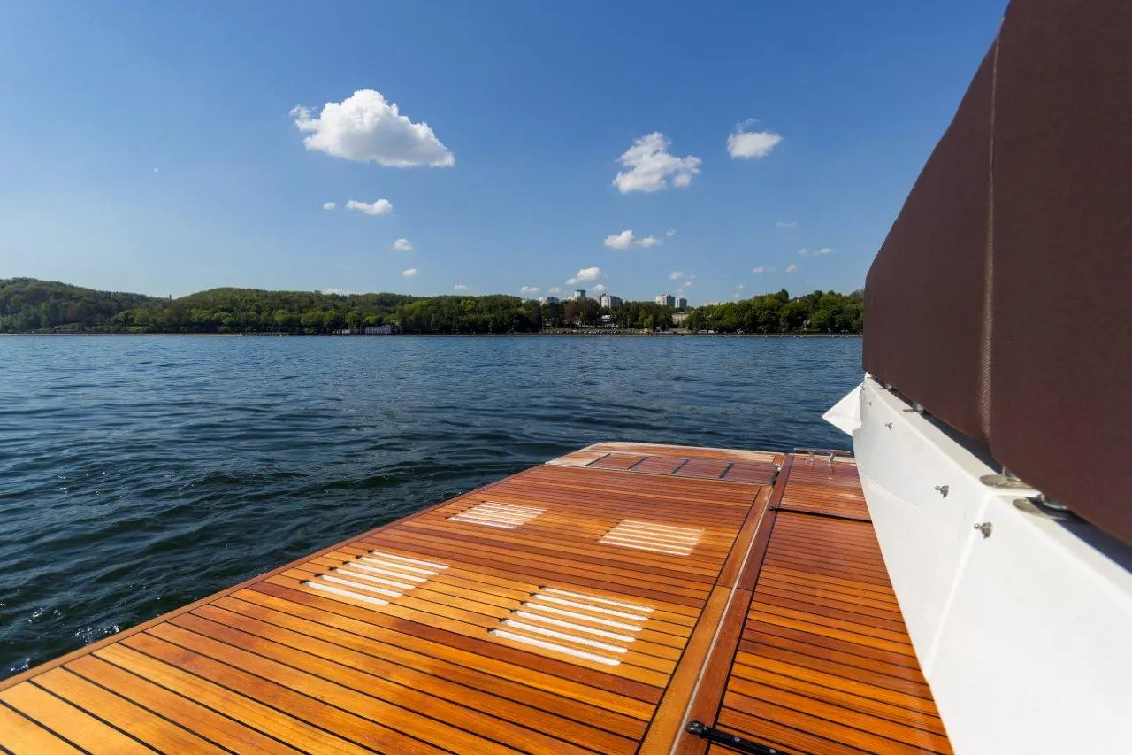 A wooden dock extends over calm water, under a clear blue sky dotted with fluffy clouds, surrounded by green hills and buildings.
