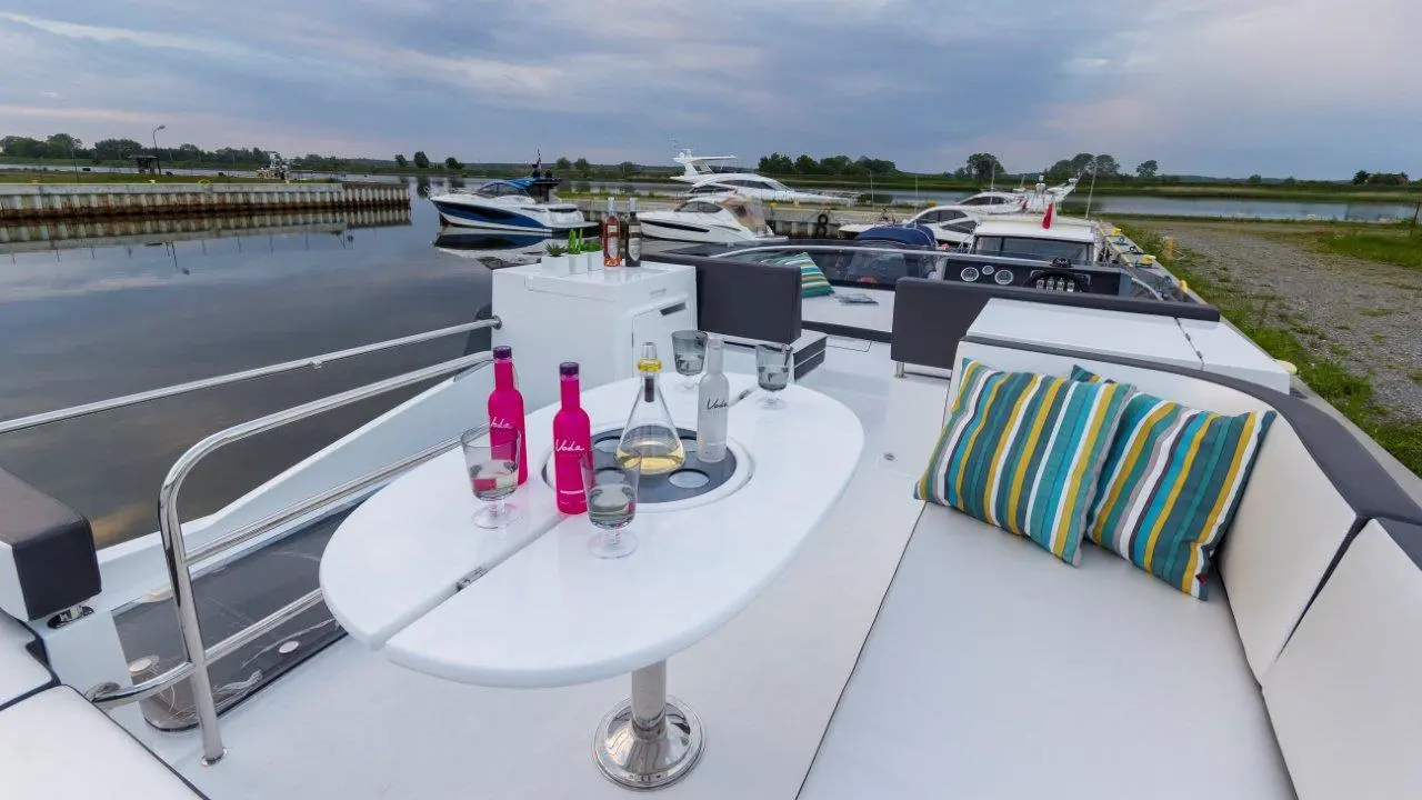 A luxury boat deck with drinks, including pink and clear bottles, and striped cushions, overlooking a marina and calm waters.