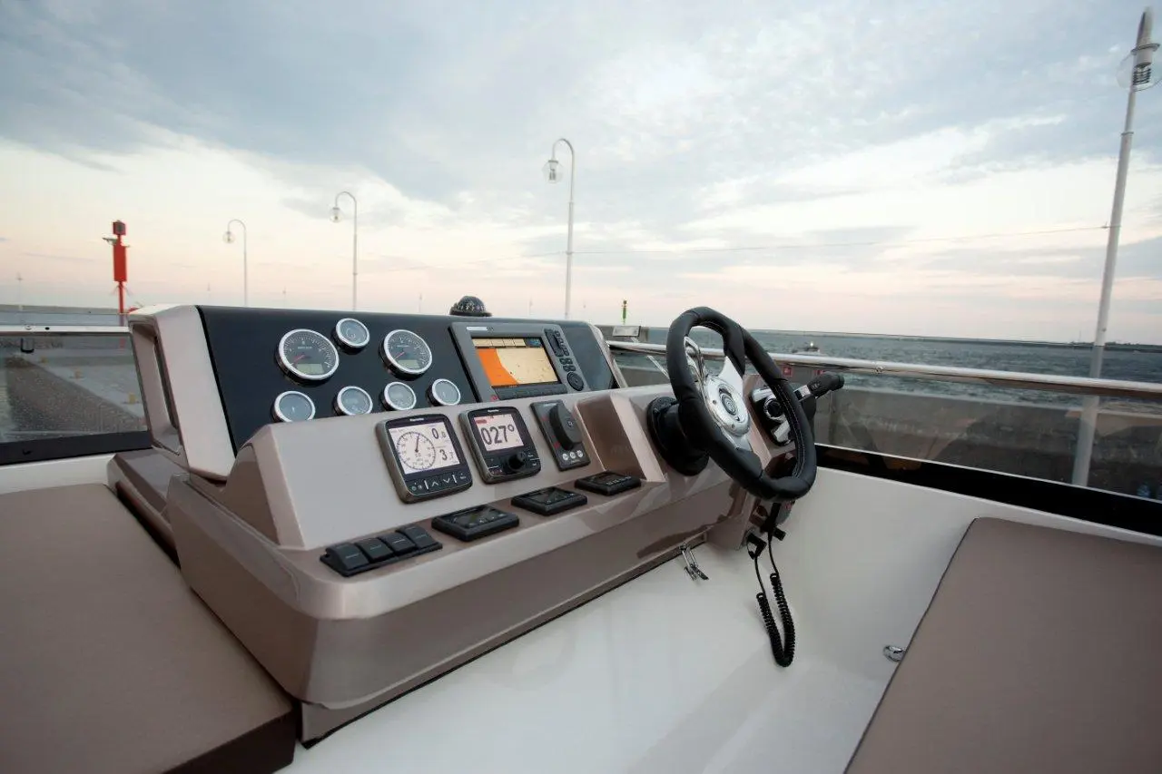 Boat cockpit with navigation instruments, gauges, and steering wheel, overlooking a calm waterway at sunset.