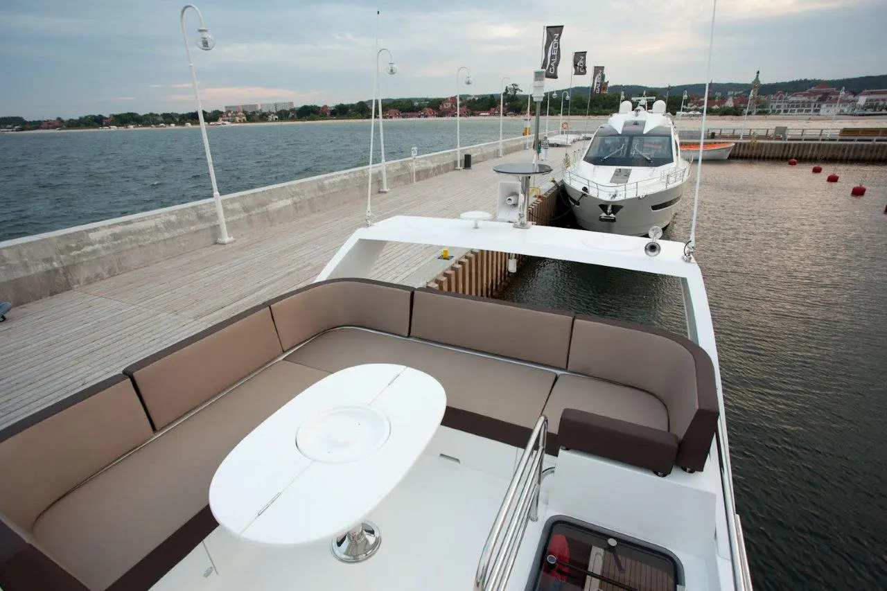 A luxury boat deck features a brown and beige sofa, a white table, and views of the waterfront and nearby docked boats.