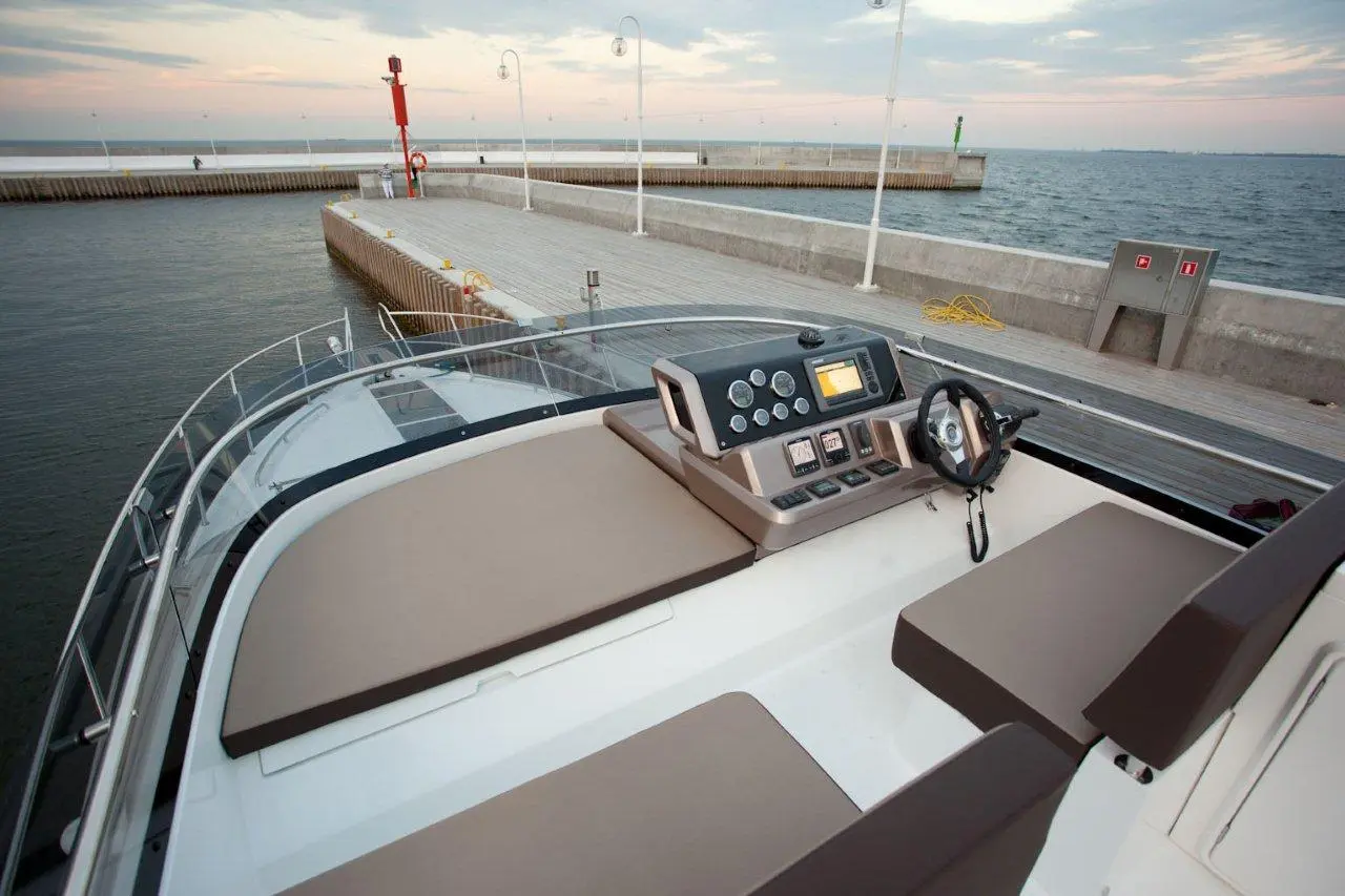 View from the helm of a luxury yacht, showcasing controls, canvas seating, and a marina with a lighthouse in the background.