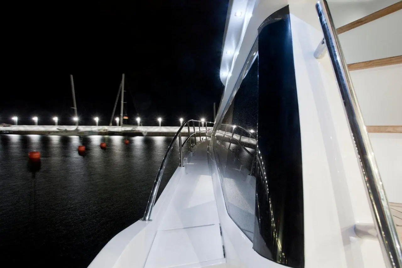 Night view from a yacht, showcasing the sleek white deck, railing, and illuminated marina with floating buoys.