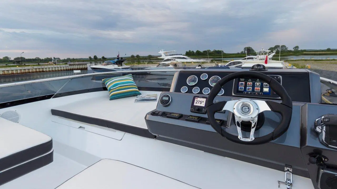 Luxury yacht cockpit featuring a steering wheel, gauges, and a touchscreen display with nearby striped cushion and boats in the background.