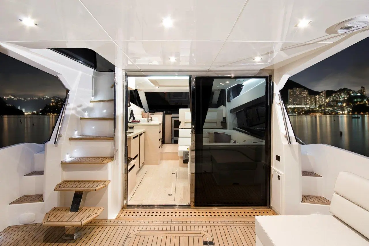Modern yacht interior at night, featuring wooden stairs, kitchen area, and views of city lights through large windows.