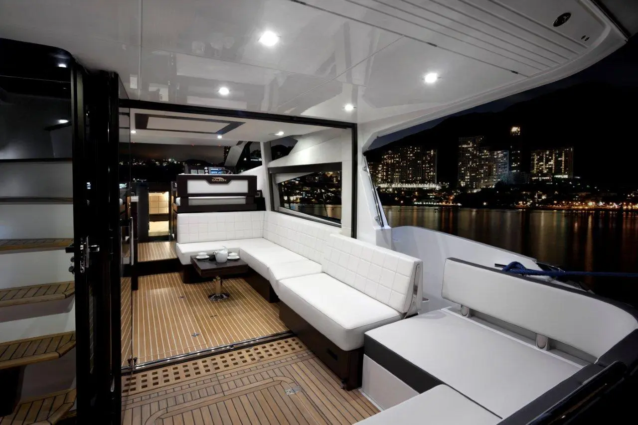 Modern yacht interior at night, featuring white seating, a wooden floor, and city skyline view through large windows.