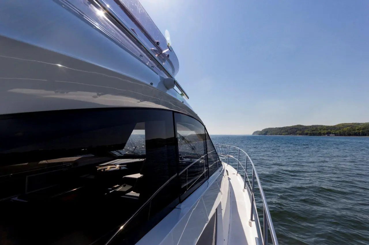 A sleek yacht's side view gliding over calm waters, with green hills visible in the distance under a clear blue sky.