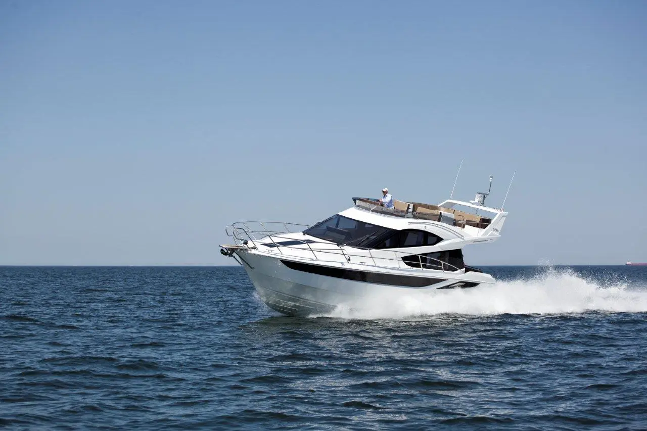 A sleek white yacht speeds across calm waters, leaving a wake, with a person steering from the upper deck.