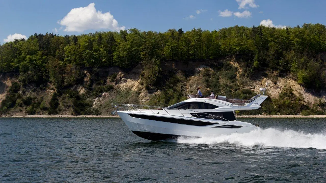 A sleek white motor yacht glides across clear water near a wooded shoreline under a sunny sky with fluffy clouds.
