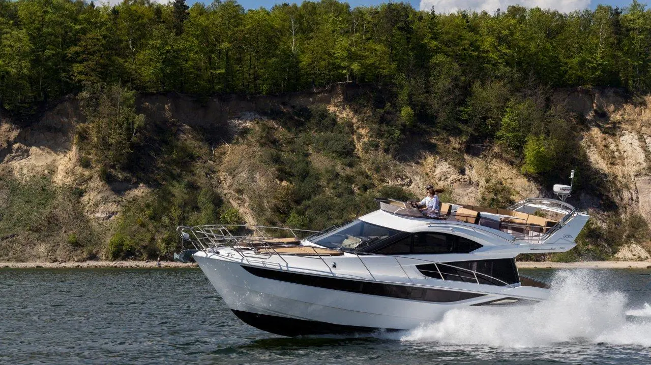 A white motor yacht speeds through calm waters, with a person at the helm, green forest in the background.