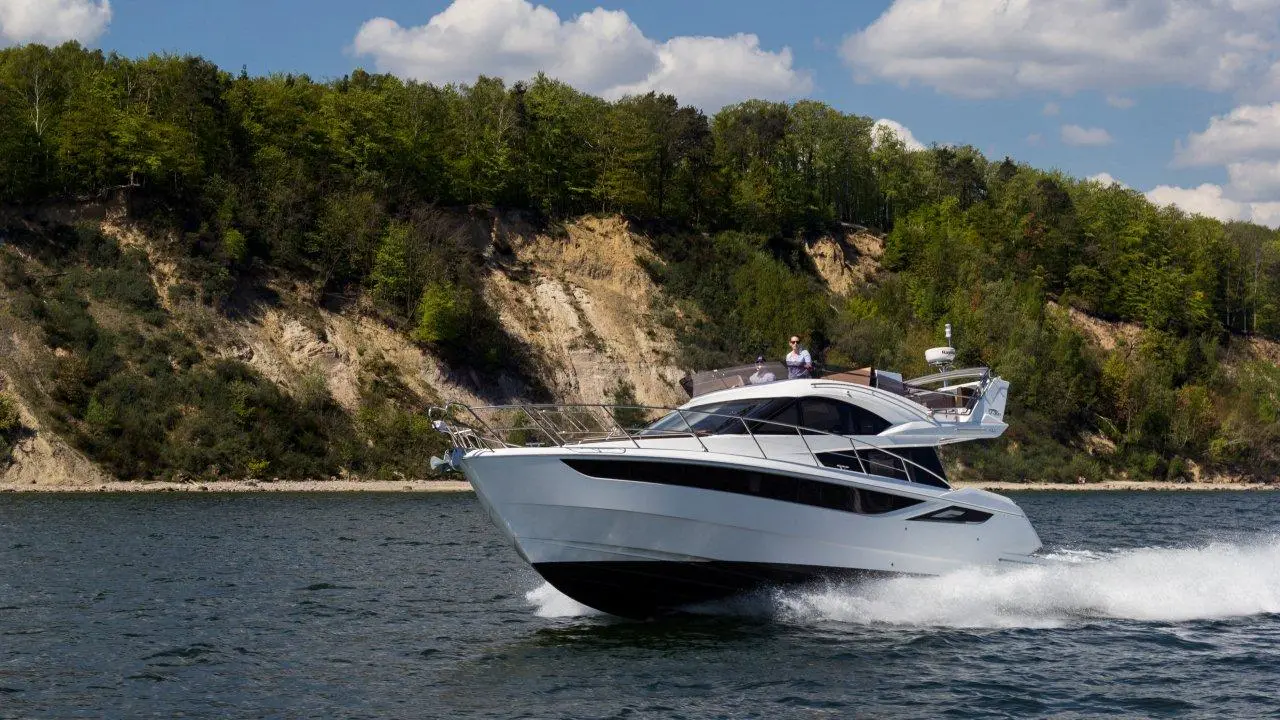A sleek white yacht cruising on calm water near a tree-lined shoreline under a blue sky with fluffy clouds.