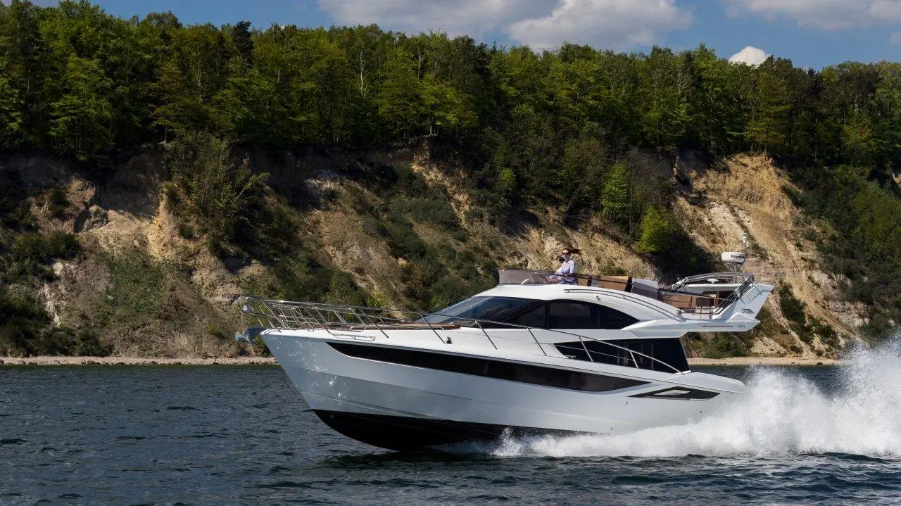 A sleek white yacht speeds across the water near a tree-lined shore, with a man relaxing on the deck.